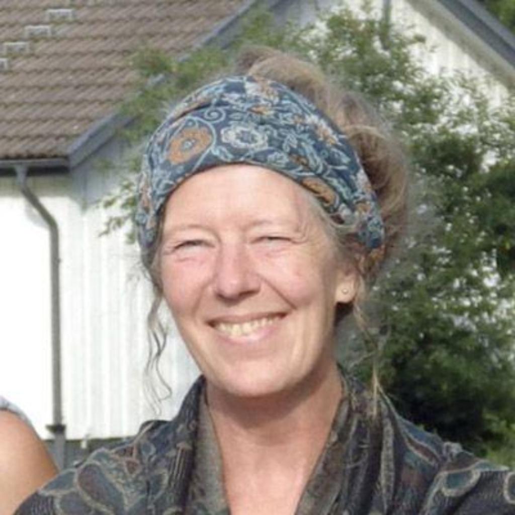 Porträt von Anna Giesler: Eine lächelnde Frau mit gemustertem Stirnband und locker hochgesteckten Haaren steht draußen vor einem Haus und grünen Bäumen